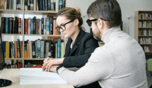 A man with low vision and a woman wearing reading glasses sit at a table in a library. A book is on the table in front of them. The woman is assisting the man to read braile.