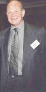 John Finch smiling and wearing a black suit with a grey shirt, a black tie and and white name badge.