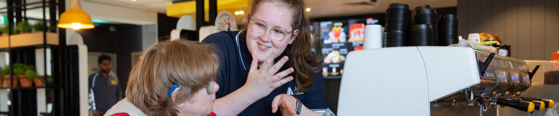 Two people inside a McCafe. They are at the counter ordering. One person is using Auslan to communicate.