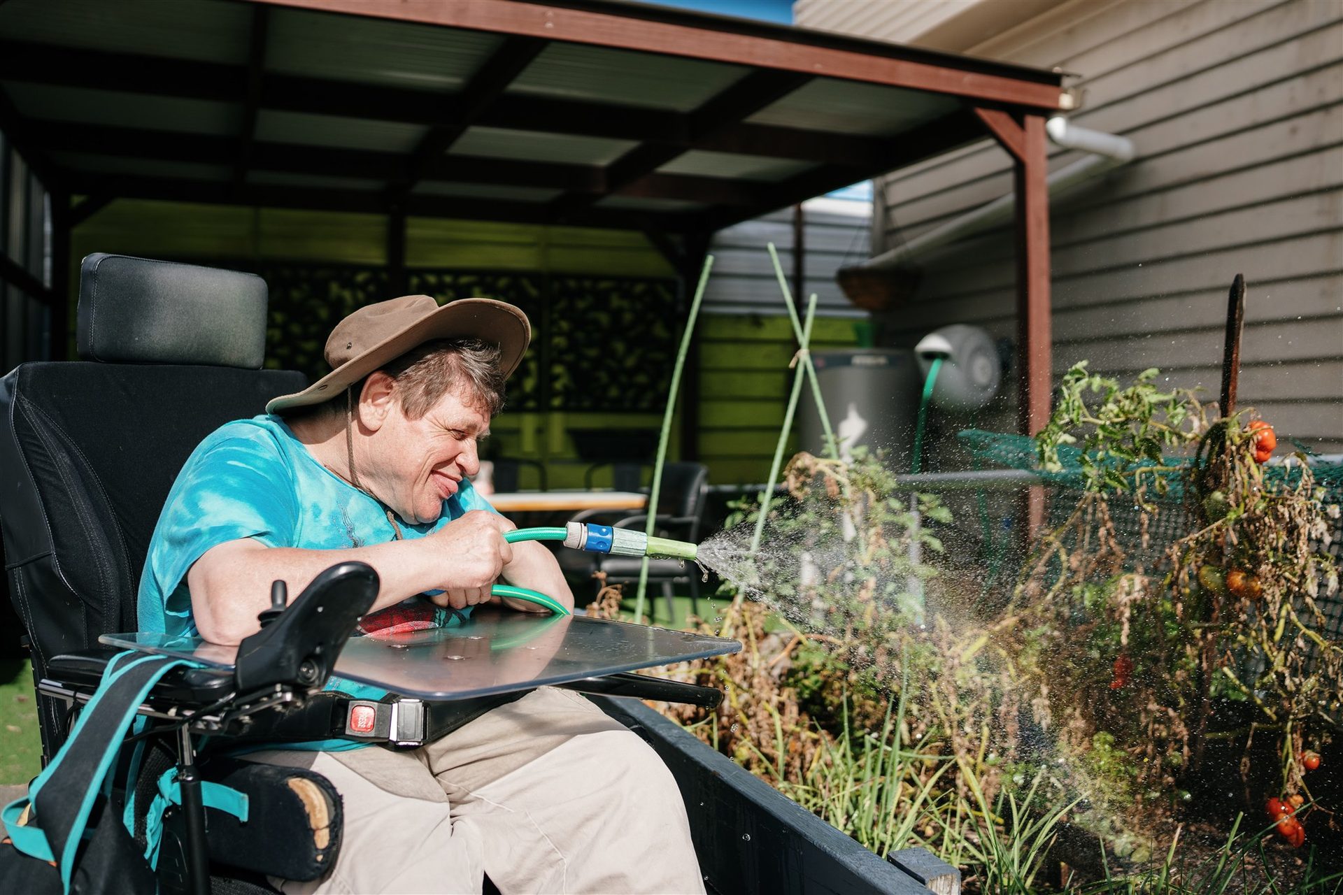 Able Australia client enjoying gardening outdoors, smiling as he waters plants from his wheelchair under a sheltered patio.