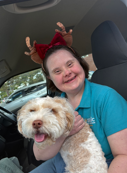 A photo of Mary Rose smiling and holding a white and brown dog. She is wearing a festive headband that has antlers and a red bow.