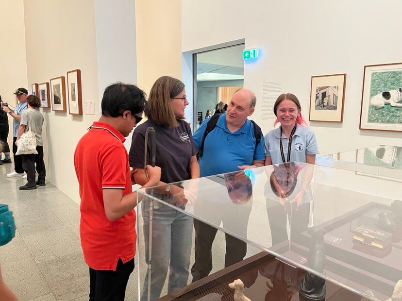A photo is of Alex, Mark, a Comm Guide and a Museum staff member facing a glass display cabinet at the exhibit.