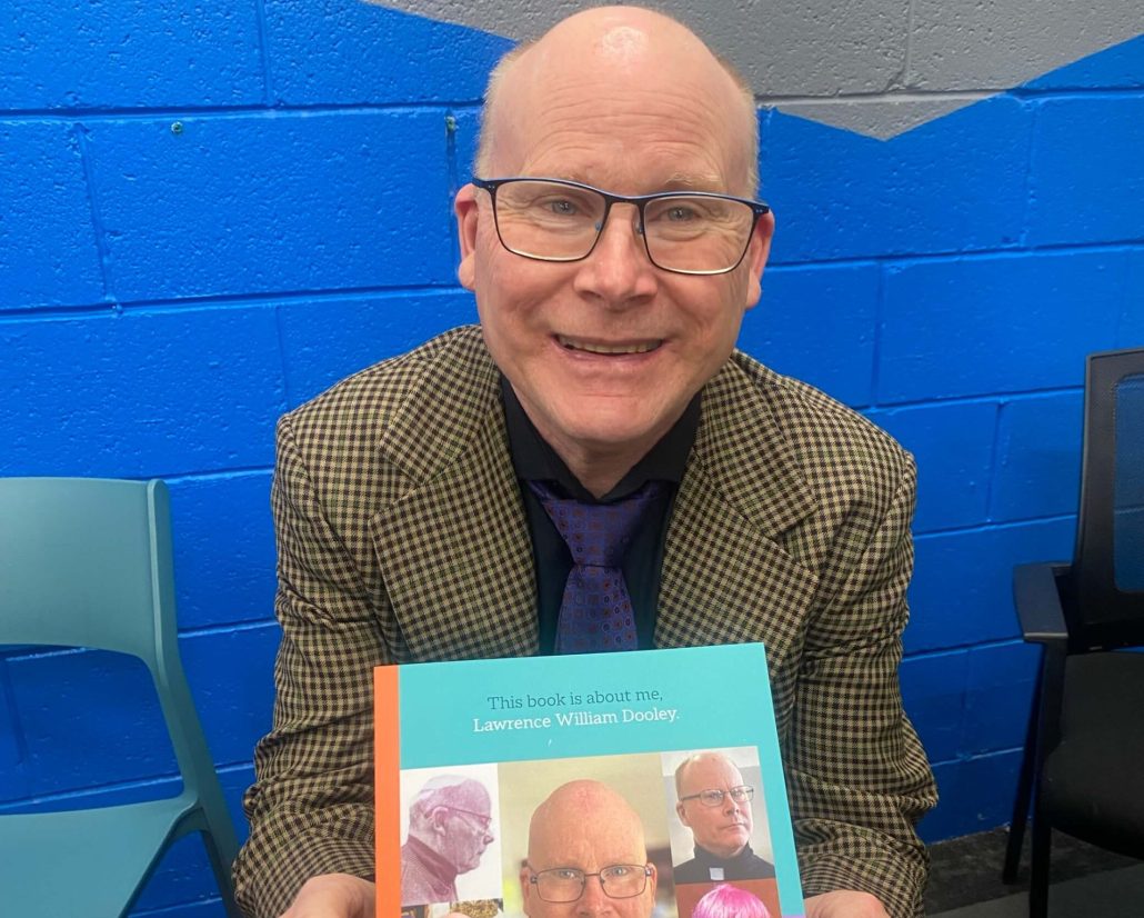 A photo of Lawrence sitting down holding his book 'This book is about me' smiling and he is wearing glasses.