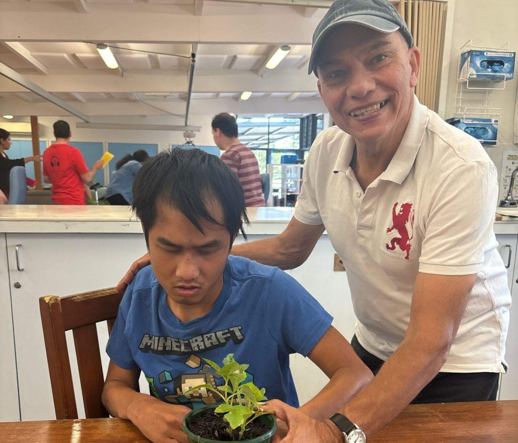A photo of Samuel seated in front of his potted plant. Next to him stands David who is smiling. Samuel is wearing a blue Minecraft t-shirt.