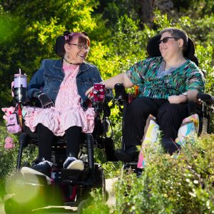 Margie and Rachael sit next to each other in their wheelchairs, surrounded by green bushes. They are looking at each other and smiling.