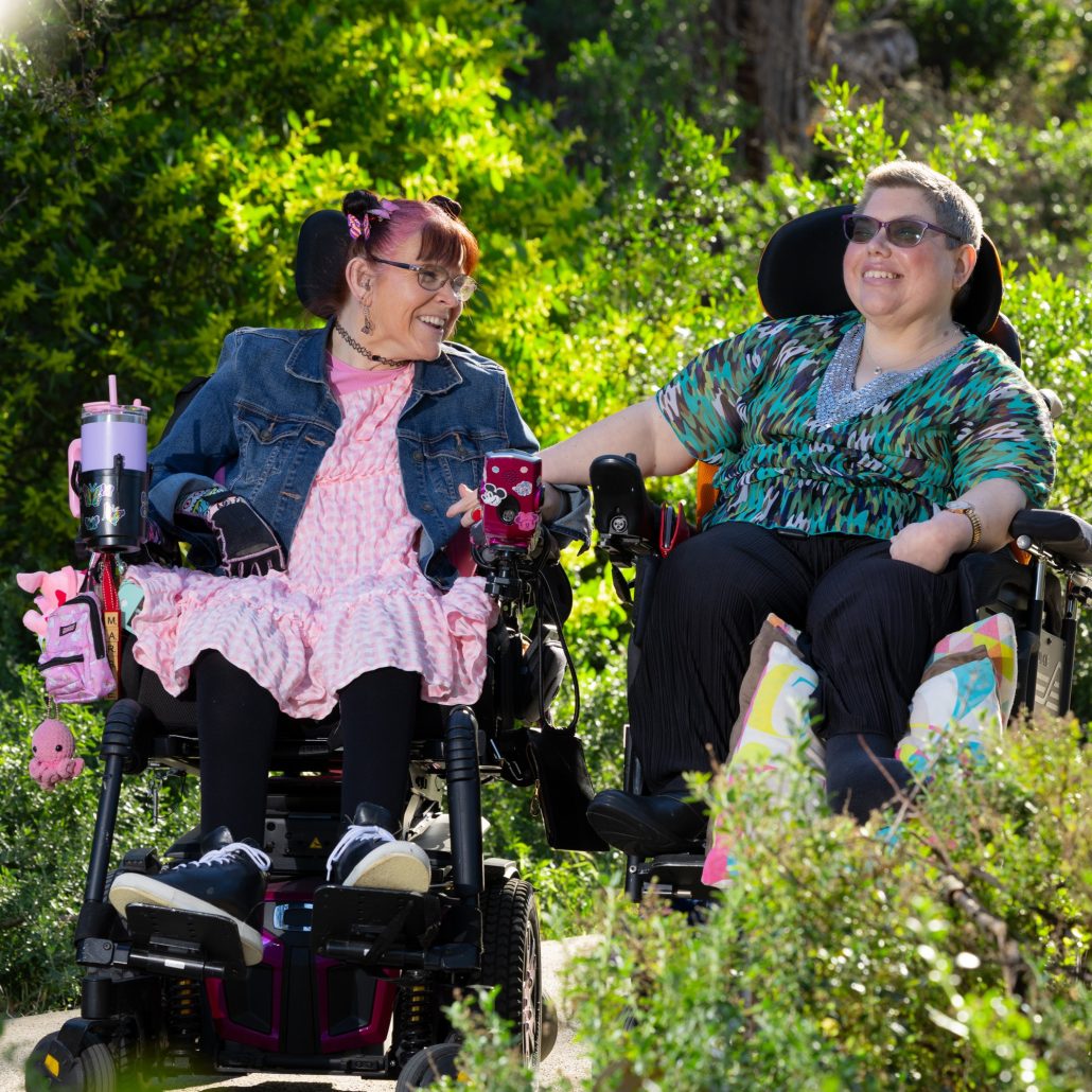 Margie and Rachel sitting in their wheelchairs smiling at eachother in the beautiful garden with the sun shining on them.