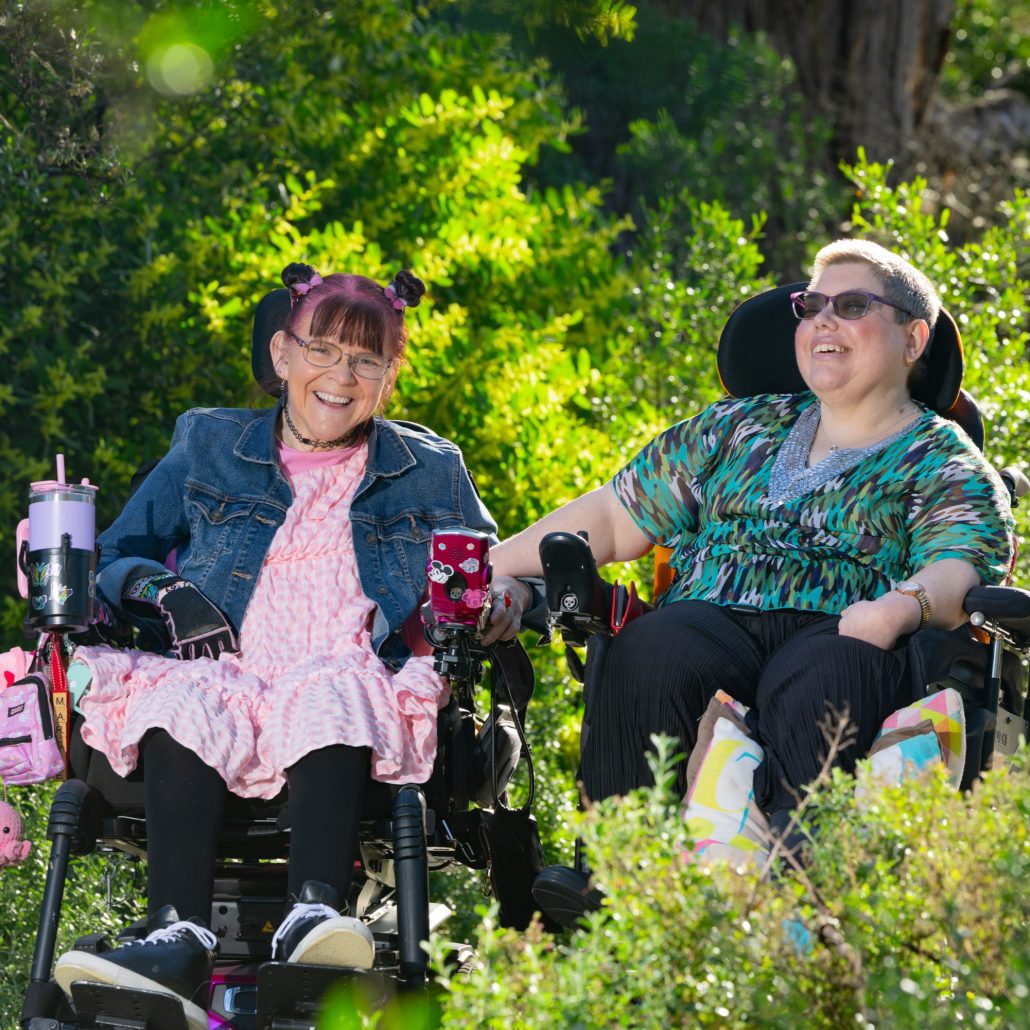 Margie and Rachel sitting in their wheelchairs smiling at eachother in the beautiful garden with the sun shining on them.