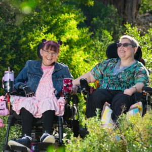 Margie and Rachel sitting in their wheelchairs smiling at eachother in the beautiful garden with the sun shining on them.