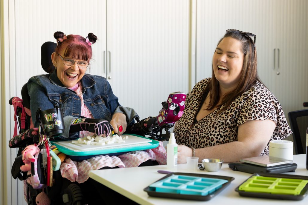 Two women sitting at a table doing arts and crafts. On the left is a woman with a big smile on her face wearing a denim jacket and her hair in two side buns she is sitting her wheelchair to the right is a woman sitting and smiling as she engages with the other woman. She has brown hair and wearing a leopard tshirt.